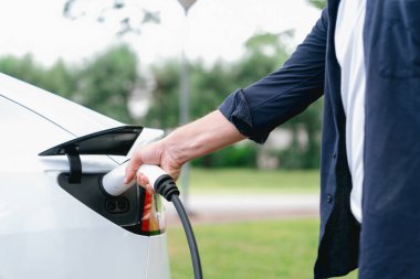 Açık yeşil şehir parkı manzaralı EV şarj istasyonundan EV elektrikli araçları şarj eden adam. Çevre dostu şehir ulaşımı ve çevre dostu EV araba seyahati. Yükselt