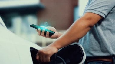 Modern iş adamı akıllı telefonun 3D hologramından batarya durumunu kontrol ederken EV 'si şarj olan enerjisini kontrol ediyor. Gelecekteki EV otomotiv teknolojisinin yenilikçi teknolojik gelişimi. İnceleyin