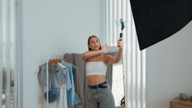Genç sosyal medyum kadın moda videosu yapıyor. Blogcu kameraya selfie çubuğu ve ışık halkası kullanarak gülümserken, aynı zamanda ikna edici çevrimiçi kıyafetlerin izleyicilere veya takipçilere vlog satmasını sağlıyor. Düzenle