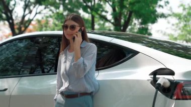 Doğal orman ya da milli parktaki araba yolculuğu sırasında elektrikli araba pili şarj ederken kadın telefonda konuşuyor. Tatil ve tatil boyunca çevre dostu seyahat. Yükselt