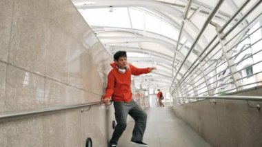 Yetenekli Asyalı break dansçı B-boy step dansını dar koridorda gösterdi. Profesyonel enerjik hippi, kameraya kendinden emin bir şekilde bakarken kulaklık takıyor ve yürüyor. Açık hava sporu 2024. Yumuşak bir şekilde..