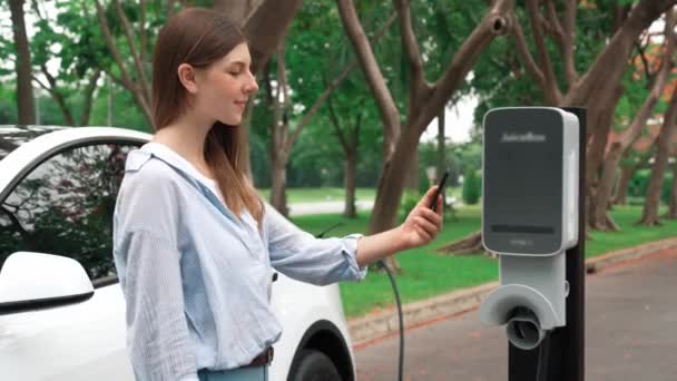 国立公園や夏の森での休暇旅行中にEv充電ステーションから電気自動車バッテリーを充電するためにスマートフォンオンラインバンキングアプリケーションを使用している女性 エクスアルト — ストック動画