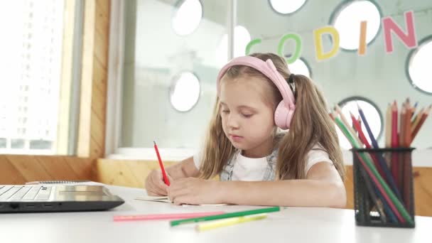 可爱的女孩一边做功课 一边在网上学习 同时听老师讲课 学生们穿着耳机 笔记本电脑和彩色铅笔写作 在纸上工作 专注于笔记本电脑 Erudition — 图库视频影像