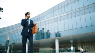 Akıllı iş adamı binada yürürken telefonla konuşuyor. İş planı, pazarlama stratejisi ya da çalışma hakkında konuşmak için akıllı telefonla konuşurken sokakta yürüyen mutlu bir yönetici. Sevinçli..