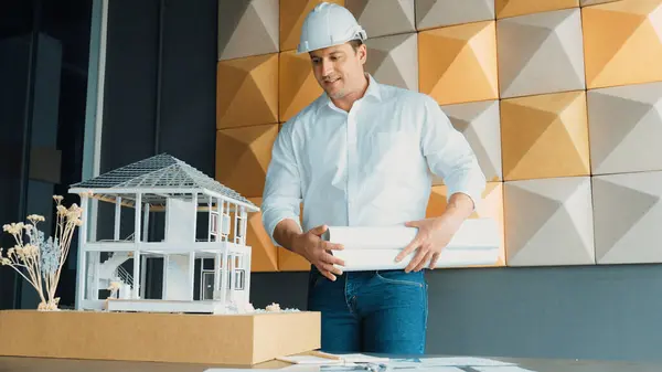 Portrait of professional project manager holding project plan with house model at table. Professional architect engineer wearing safety helmet while standing posing with confident. Manipulator.
