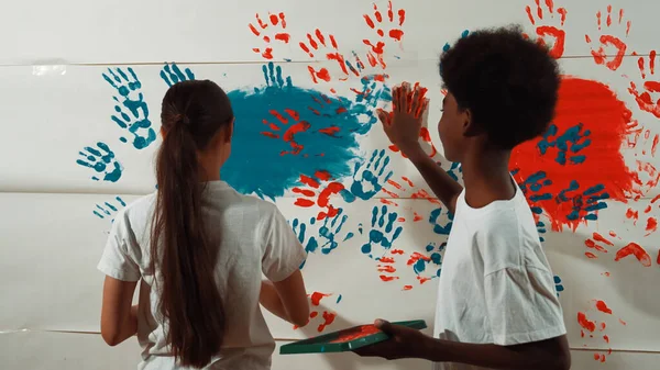 Diverse happy highschool girl and african boy paint the wall with water color in red and blue or contrast color by using their hands in art lesson. Colorful. Creative activity concept. Edification.