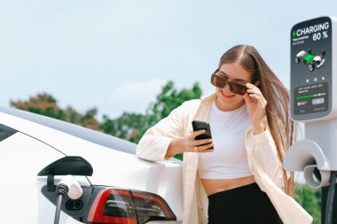 Genç bir kadın EV otomobil şarj istasyonu Green City Park 'ta elektrik ödemek için akıllı telefonu kullanıyor. Modern çevre ve EV araçlı sürdürülebilir şehir yaşam tarzı. Yararlı