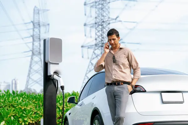 在与电网塔相连的充电站充电时 人们一边打电话一边给电动汽车电池充电 一边谈论环保汽车的应用 — 图库照片