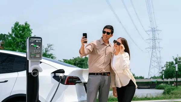 在与电网塔电相连的充电站为电动汽车充电时 夫妇用智能手机支付电费 以促进环保汽车的使用 — 图库照片