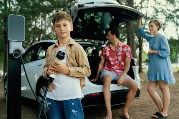Evチャージャーを抱えている小さな少年が 家族と一緒にバックグラウンドのトランクに座ってカメラを指さした 環境に優しい車のコンセプトのための代替エネルギー充電ステーションとロード旅行 パーペチュアル — ストック写真