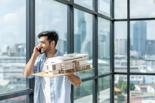 Handsome skilled architect engineer holds architectural model while inspect house model. Professional interior designer checking house construction while standing near window with city view. Tracery.
