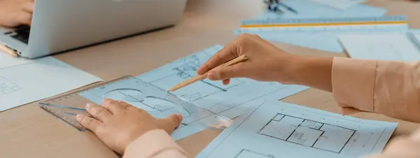 Professional architect drawing blueprint during meeting at modern architectural office on table with architectural document and stationary scatter around. Blurring background. Closeup. Delineation