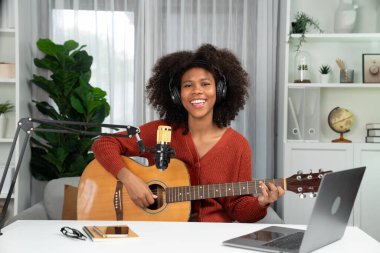 Genç Afrikalı Amerikalı müzisyenin sunucu kanalı gitar çalıyor, şarkı söylüyor, stüdyoda dizüstü bilgisayardan yayın yapıyor. Kulaklık dekorasyonu ve mikrofon kaydı. Tatlandırıcı.