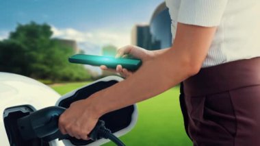 Modern kadın EV akü durum hologramını akıllı telefon uygulamasından kontrol ederken EV otomobilini EV 'nin şarj istasyonundan ağaç ile şarj ediyor. Geleceğin yenilikçi enerji sürdürülebilirliği. İnceleyin