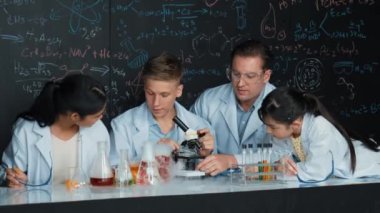 Çok kültürlü bir öğrenci mikroskop altında incelerken güzel bir kız da numuneyi inceliyor. BTMM sınıfında karatahta bilim teorisi deneyi yapan liseli çocuklara bakan öğretmen. Öğretim