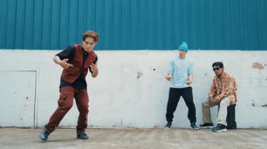 Genç Asyalı dansçı hip hop tarzında dans ediyor ve çok kültürlü arkadaşları duvarın arkasında tezahürat yapıyor. Çekici yakışıklı adam mavi arka planda kollarını uzatıyor. Açık hava sporu 2024 hiphop..