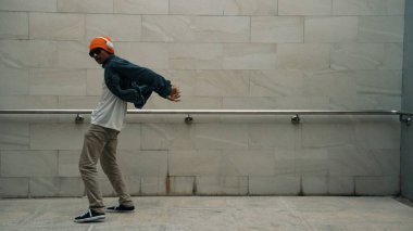 Asyalı akıllı hipster, kulaklık ve şık kıyafetler giyip hip hop şarkıları dinlerken gri arka planlı enerjik break dans gösterisi yapıyor. Açık hava sporu 2024. Hip hop hareketi. Yumuşak bir şekilde..