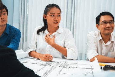 Mimari proje üzerinde birlikte çalışan çeşitli inşaat mühendisi ve müvekkil grupları, inşaat planını gözden geçirip toplantı masasında plan oluşturuyorlar. Tedbirli