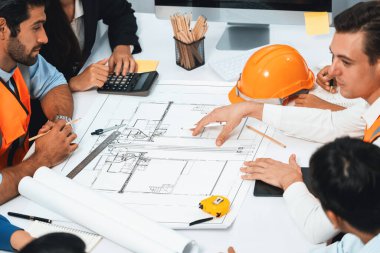 Mimari proje üzerinde birlikte çalışan çeşitli inşaat mühendisi ve müvekkil grupları, inşaat planını gözden geçirip toplantı masasında plan oluşturuyorlar. Tedbirli