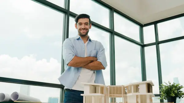 Portrait of architect engineer in casual outfit smile at camera while crossing arms. Businessman looking at camera and standing with arms folded near house model, architectural model. Tracery