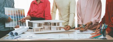 Profesyonel mimar takımının ev rengi seçimi hakkında beyin fırtınası yaparken, mimar da plan ve ev modelinin olduğu masadaki taslak belgeye yazı yazıyor. Değiştirilmiş.