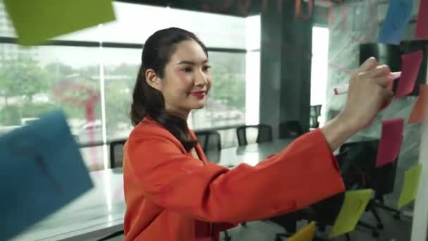 年轻美丽的女商人在玻璃板上书写营销理念的肖像 亚洲项目经理运用五彩缤纷的贴纸来分享营销策略 头脑风暴启动项目 机械手 — 图库视频影像