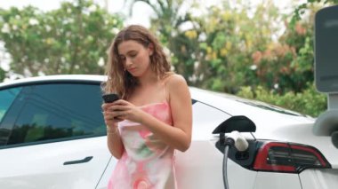 Genç bir kadın doğadaki EV şarj istasyonunda elektrik için akıllı telefon kullanıyor. Modern çevre ve EV araçlı sürdürülebilir otomobil taşıma yaşam tarzı. Titiz
