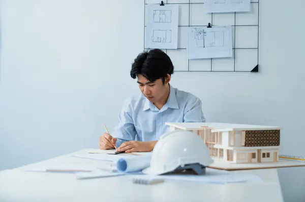 Closeup portrait image of architect engineer writing, drawing, taking a note about house model structure with safety helmet blueprint, placed on table. Business creative design concept. Immaculate.