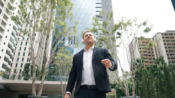 Business people wear headphone to listen relaxing music while walking workplace at green city. Low angle camera of manager move to song while go to meeting surround with tree and skyscraper. Urbane.