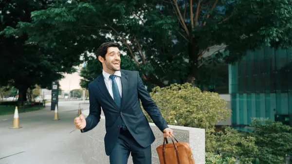 Smart business man walking along the road while holding bag at park. Professional project manager smiling while traveling in lively mood. Happy investor getting a promotion. Business dance Exultant.