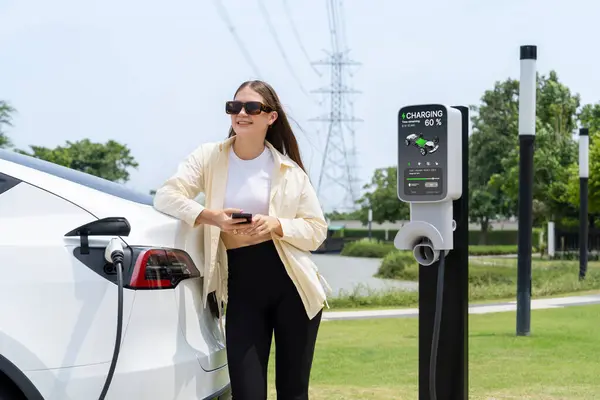 若い女性は公共電気自動車の充電ステーショングリーンシティパークで電気を支払うためにスマートフォンを使用します Ev車両による近代的な環境と持続可能な都市ライフスタイル エクスペディエント — ストック写真