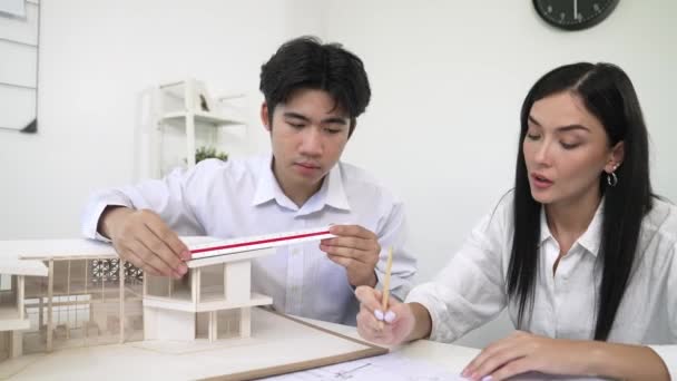 专业的亚洲建筑师工程师测量房屋模型的长度 与高加索同事一起使用现代办公室的笔记本电脑分析数据写下蓝图 纯洁无瑕 — 图库视频影像