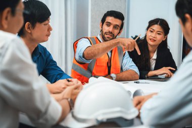 Mimari proje üzerinde birlikte çalışan çeşitli inşaat mühendisi ve müvekkil grupları, inşaat planını gözden geçirip toplantı masasında plan oluşturuyorlar. Tedbirli