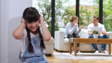 Stresli ve mutsuz genç kız köşeye kıvrılıp kulaklarını kapatarak arka planda ebeveyninin tartışmasını engelliyor. Evde aile içi şiddet ve travmatik çocukluk depresyona dönüşür. Eşzamanlama