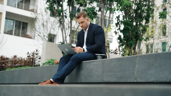 Skilled male leader working on his phone sending report while using laptop analyze investment data at green city. Happy investor looking marketing plan while sitting at park or eco urban city. Urbane.