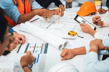 Mimari proje üzerinde birlikte çalışan çeşitli inşaat mühendisi ve müvekkil grupları, inşaat planını gözden geçirip toplantı masasında plan oluşturuyorlar. Tedbirli