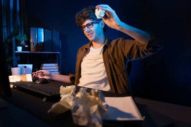 Aşırı çalışan projeyle uğraşan stresli yaratıcı yönetici buruşmuş kağıtların etrafını sararken gece vakti modern ofisteki gündelik gömlekle dizüstü bilgisayar ekranına e-posta göndermeyi bekliyor. Toplayıcı.