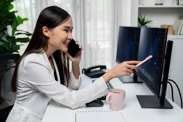 Young beautiful Asian businesswoman calling to broker, pointing with pen in increased exchange stock market on pc to invest high profit at modern office, investing high profit in wealth. Stratagem.