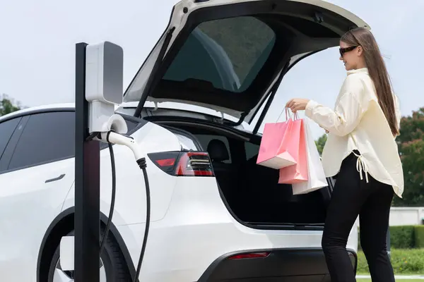 グリーンシティパークで充電ステーションからEvカーバッテリーを充電するショッピングバッグを持っている若い女性 現代の女性は 環境に優しい電気自動車旅行のライフスタイルで買い物をします エクスペディエント — ストック写真