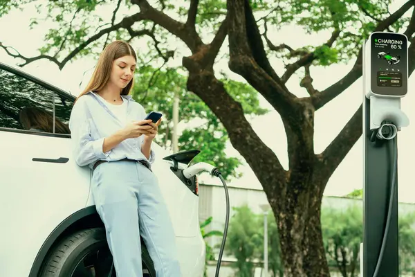 若い女性はEv充電ステーションからEv電気自動車バッテリーを充電し スマートフォンオンラインバンキングを使用して市立公園で電気を支払います Ev車を搭載したエコフレンドリーな車両 エクスアルト — ストック写真