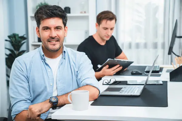 Profile of smiling freelancer or businessman looking at camera with coffee cup while colleague working on laptop with paperwork at modern office. Theme of creative project at workplace. Sellable.