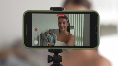 Genç sosyal medyum kadın moda videosu yapıyor. Blogcu kameraya selfie çubuğu ve ışık halkası kullanarak gülümserken, aynı zamanda ikna edici çevrimiçi kıyafetlerin izleyicilere veya takipçilere vlog satmasını sağlıyor. Düzenle
