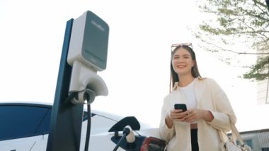 Ağır çekimde genç bir kadın, alışveriş merkezinin otoparkındaki şarj istasyonundan şarj olan EV 'nin pilini şarj ederken akıllı telefondan akü durumunu kontrol ediyor. Modern kadın elektrikli araçla alışverişe gider. Yararlı