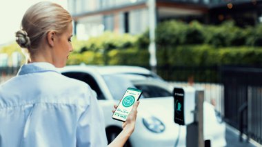 İş kadını, çevre yeşil şehir parkındaki şarj istasyonundan şarj olurken akıllı telefon ekranından EV batarya durum mobil uygulamasını kontrol ediyor. Elektrikli arabanın gelecekteki enerji sürdürülebilirliği.