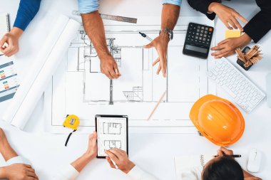Mimari proje üzerinde birlikte çalışan, inşaat planını gözden geçiren ve toplantı masasında plan oluşturan çeşitli inşaat mühendisi ve müşteri gruplarından oluşan üst düzey bir pankart. Tedbirli