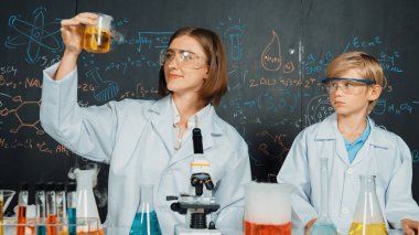 Kimyasal sıvıyı bilim laboratuarında karıştıran zeki beyaz bir öğretmen. Profesyonel eğitmen, BTMM fen sınıfında mikroskopla karatahta yazılı kimya teorisi üzerinde deney yapıyor. Etkinlik.