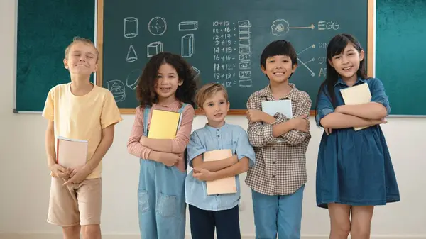 教室に立っている間カメラを見ながらカジュアルな布で多文化小学生 かわいい多様な子供のグループは 教室の黒板でノートと笑顔をカメラに保持しています ペダゴロジー — ストック写真