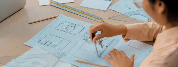 Professional architect drawing blueprint by using divider on the table with stationary and architectural document scatter around at architectural office. Closeup. Focus on hand. Delineation.
