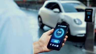 Akıllı telefon ekran bataryasını elinde tutan iş adamı akıllı EV mobil uygulama ile otomobil parkındaki şarj istasyonundan elektrik şarj eden çevre dostu EV otomobil. İnceleyin