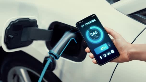 スマートEvモバイルアプリによるスマートフォンディスプレイのバッテリーステータスインターフェースを持つビジネスマンや 駐車場の充電ステーションから電気を充電する環境に優しいEvカー Peruse — ストック動画